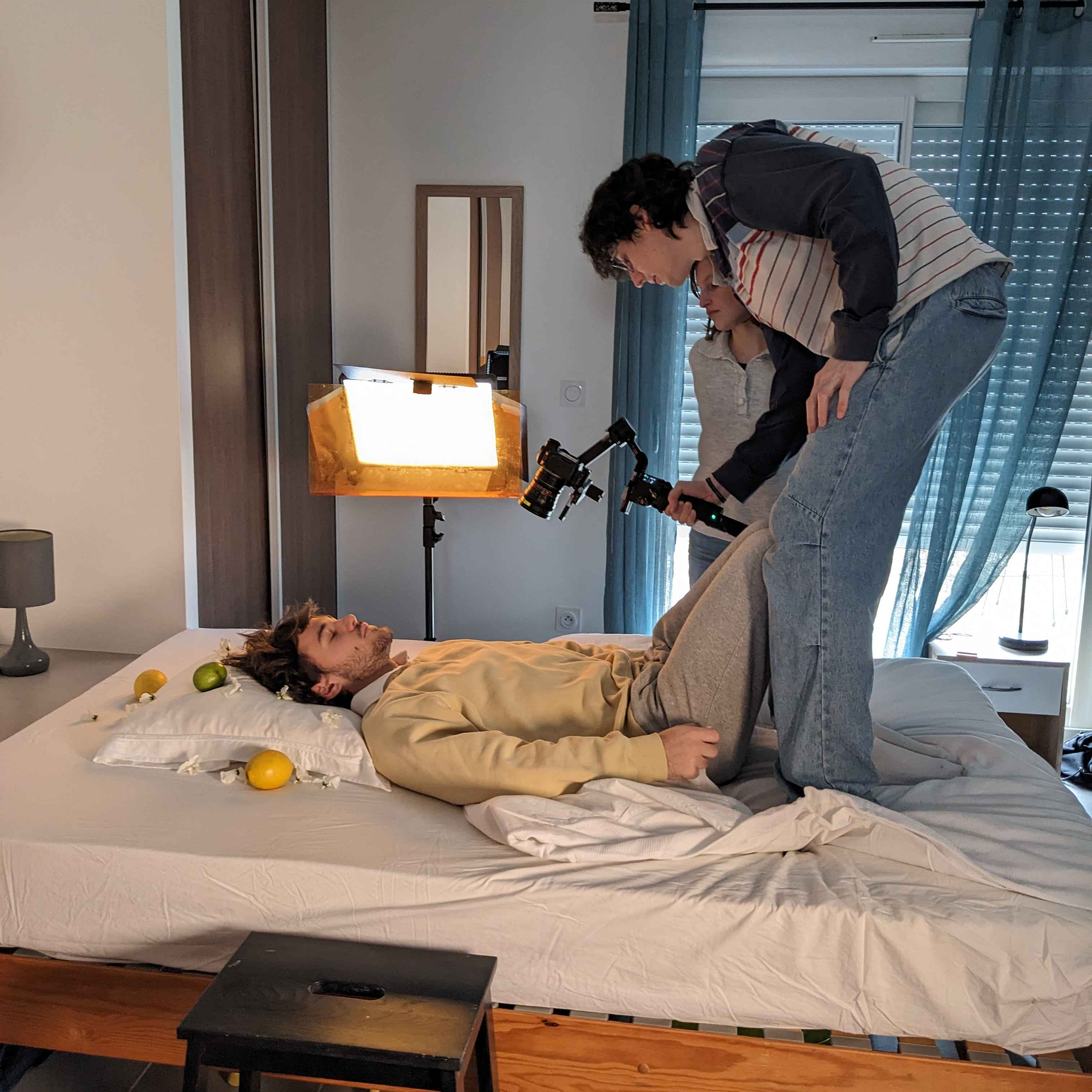 Photo de tournage : Marius debout sur le lit au-dessus de Louis pour filmer la première scène de la vidéo, Lucie derrière, supervisant.